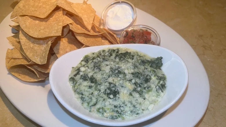 Cheesecake Factory Hot Spinach and Cheese Dip in white dish next to tortilla chips and cups of sour cream and salsa