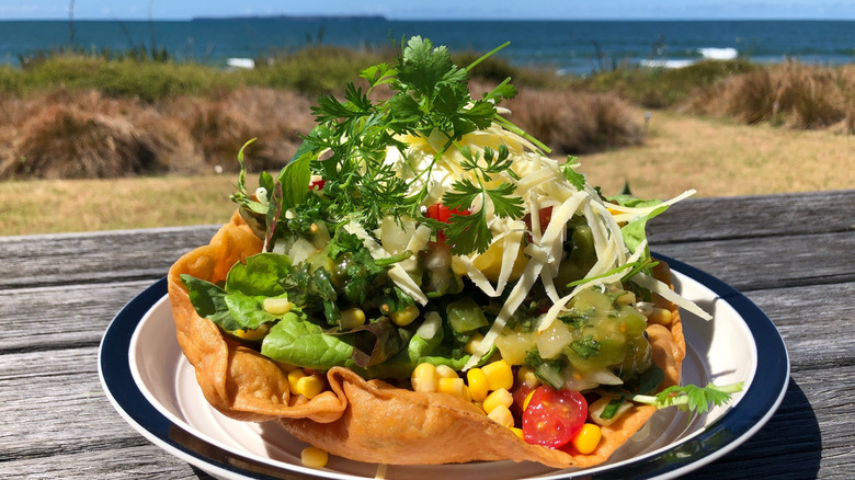 A crispy tortilla bowl loaded with salad, sitting on a plate on a picnic table outside