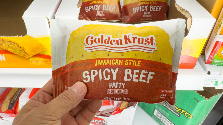 A hand holding a packaged frozen Golden Krust beef patty.
