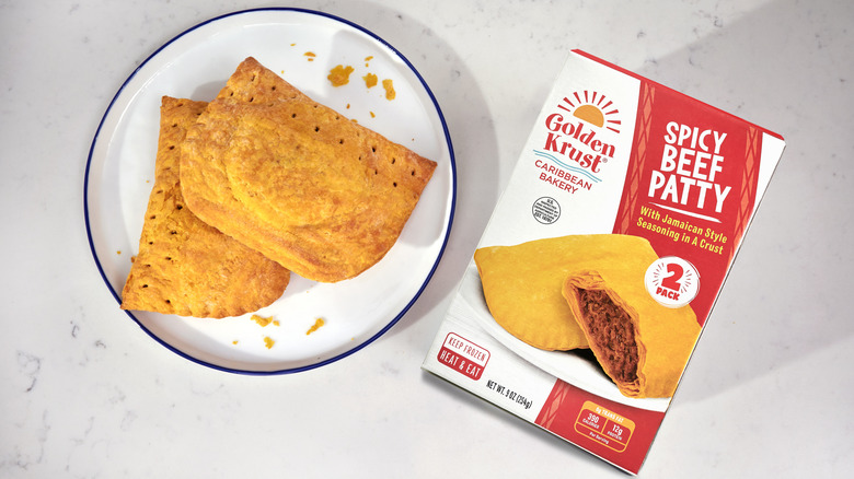 A hand holding a packaged frozen Golden Krust beef patty.