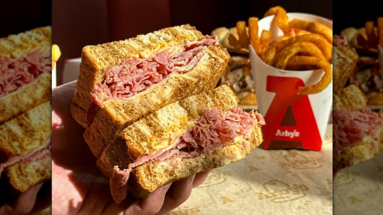 Someone holding an Arby's roast beef sandwich on thick bread with curly fries in an Arby's container in the background