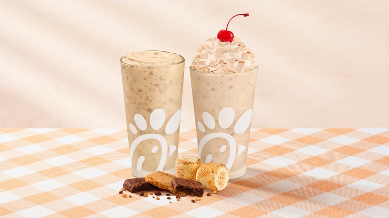 A pair of Chick-fil-A s'mores milkshakes, one with a cherry on top, sitting on a tablecloth coverage table next to some toasted marshmallows, chocolate, and graham cracker