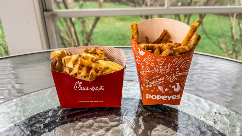 a container of chick fil a fries and popeyes fries on a glass table