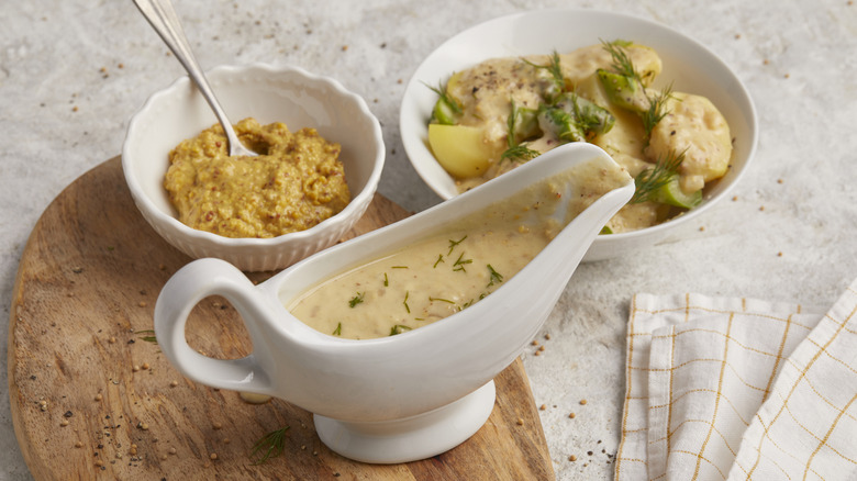 A gravy boat with Dijon sauce with a bowl of mustard and braised chicken in the background.