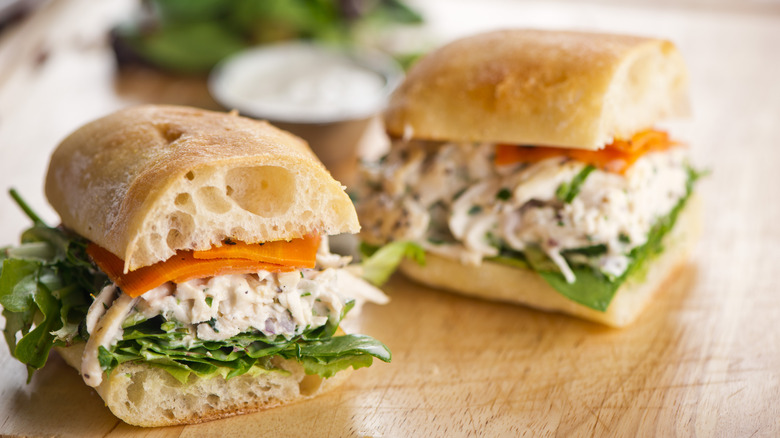 A chicken salad sandwich with lettuce and tomatoes sits on a wooden block
