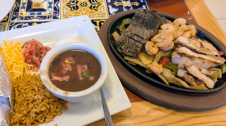 A plate of fajitas with steak, shrimp, and chicken next to another plate of rice, beans, pico, and sour cream
