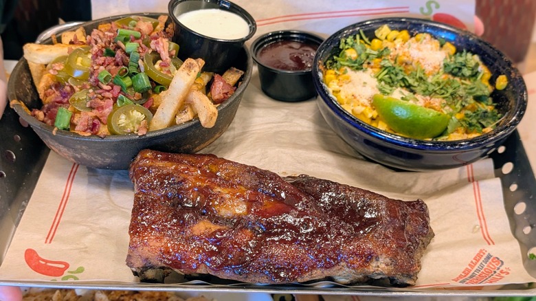 A plate of saucey ribs with bowls of bacon-topped cheesy fries and street corn salad
