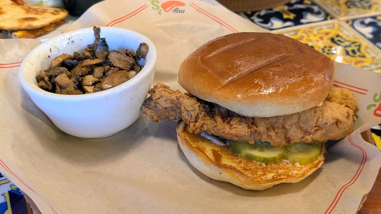A crispy chicken sandwich and a white dish of grilled mushrooms on a paper-lined plate