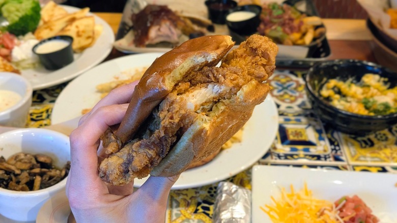 A hand holding a crispy chicken sandwich with a bite taken out with various Chili's entrees in the background