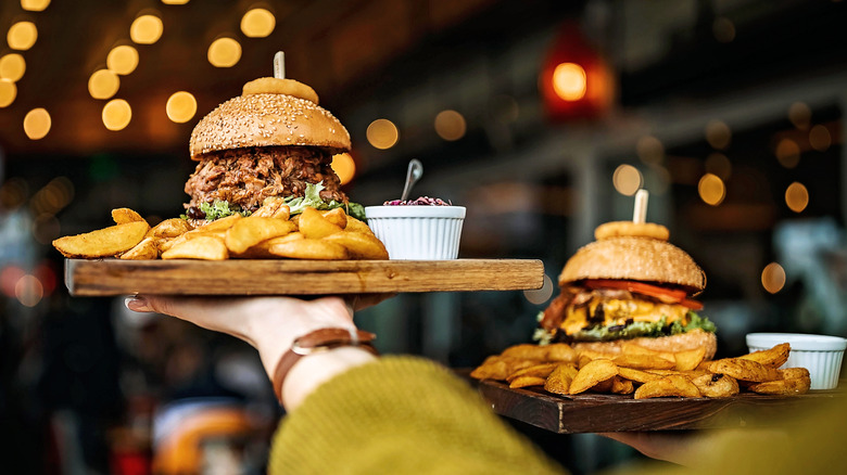 A person carrying two burgers on wooden platters, one slightly higher than the other