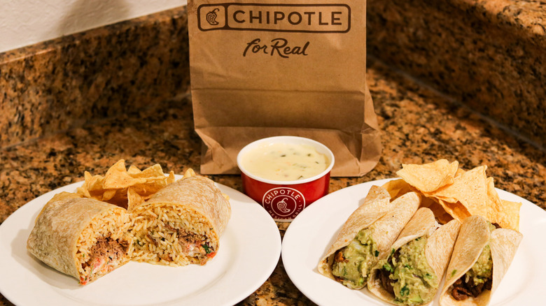 A Chipotle burrito sits on a plate next to Chipotle tacos, a cup of queso sits between, and a brown paper Chipotle bag behind them.