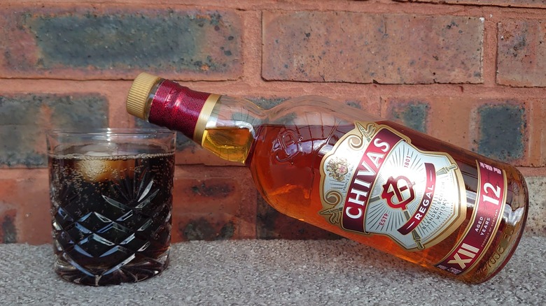 A bottle of Chivas Regal 12-year resting on a glass of whisky and cola with ice