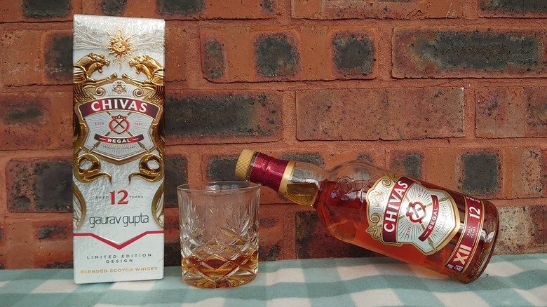 A bottle of Chivas Regal 12-year, its box and a glass shown on a red brick background