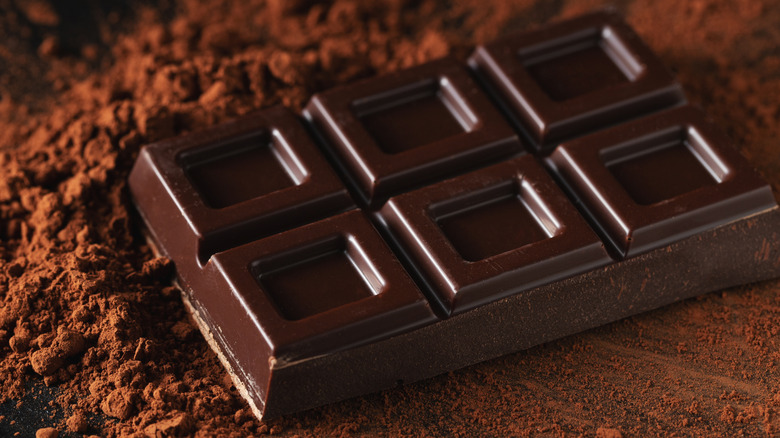 Bar of chocolate on top of pile of cocoa powder on dark surface