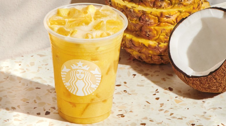A yellow Starbucks Iced Paradise drink next to a pineapple and coconut