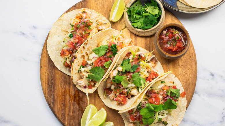 Four shrimp tacos on round wooden cutting board.