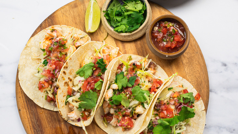 Four shrimp tacos on round wooden cutting board.