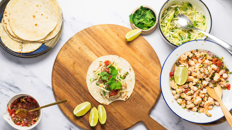 One shrimp taco on round wooden cutting board with all the components around.