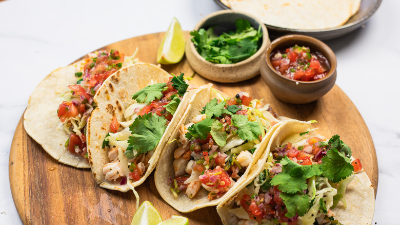 Four shrimp tacos on round wooden cutting board with tortillas in background.