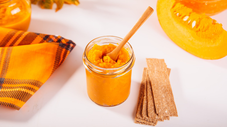 A jar of pumpkin puree with a spoon in it next to some quartered pumpkins and a cloth
