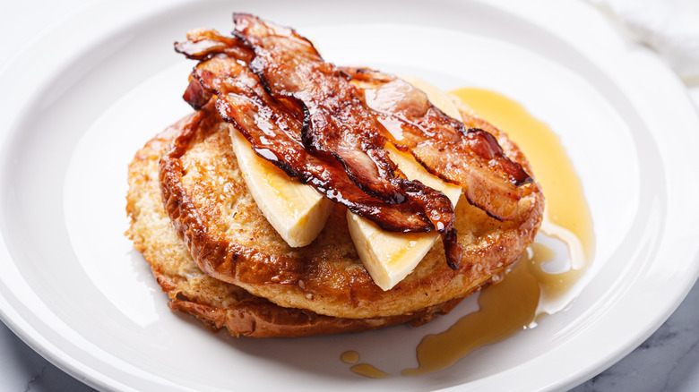 Two round slices of French toast topped with banana, bacon, and syrup.