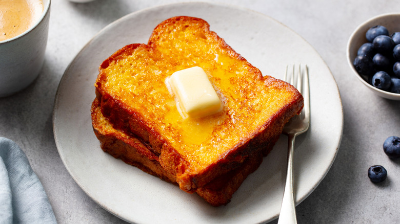 Two French toast slices with butter on white plate with fork.