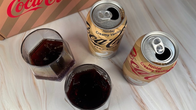 Two glasses of Coca-Cola next to two cans of Coca-Cola, branded box behind