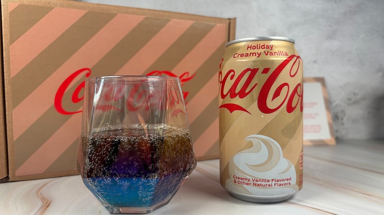 Can of Holiday Creamy Vanilla Cocal-Cola next to Coke in glass with branded box behind