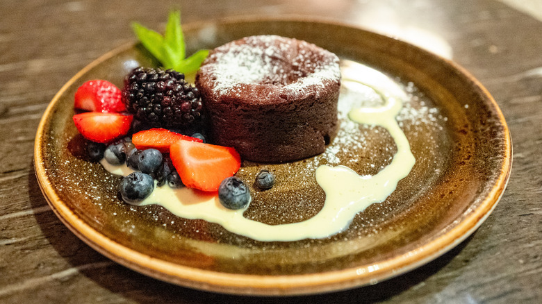 A small chocolate cake on a brown plate dusted with powdered sugar garnished with fresh berries and a white sauce