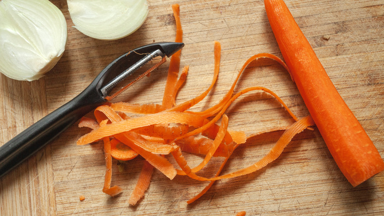 A vegetable peeler, a halved onion, a peeled carrot, and a pile of the carrot peels on a wooden cutting board