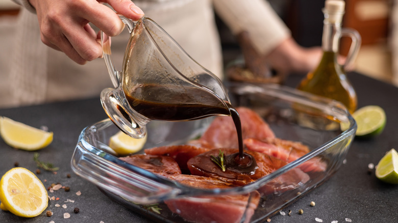 Person pouring soy sauce based marinade over meat in dish.