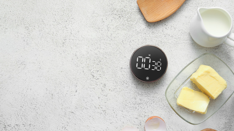 Overhead shot of kitchen timer alongside butter, milk, eggshell, and wooden spatula