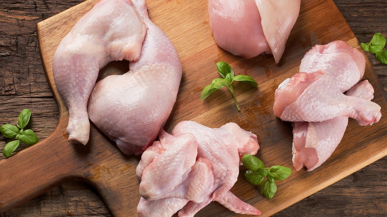 Various cuts of raw chicken on a wood cutting board with fresh sprigs of basil.