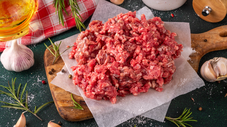 Raw ground beef of a wooden cutting board surrounded by seasonings and garlic cloves.