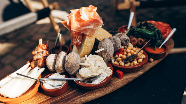 A stack of various tapas, ham, melon, bread, beans, and peppers piled on a wooden board