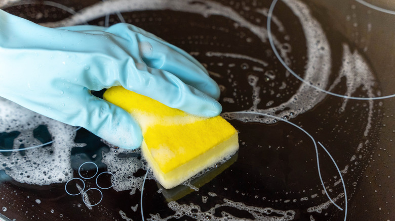 Close-up of a blue-gloved hand scrubbing a glass cooktop with soapy water and a soft yellow sponge