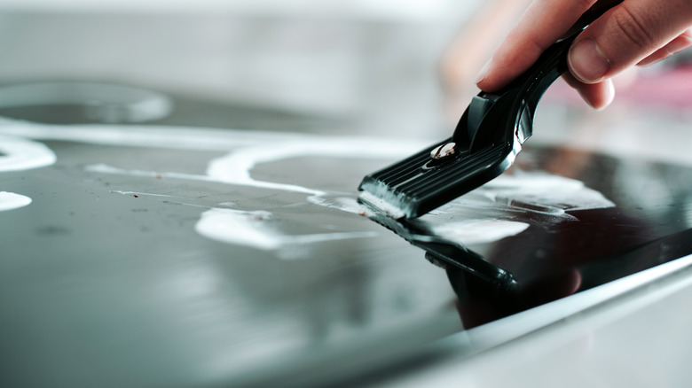 Hand scraping a glass cooktop with a single-edge razor blade in a plastic holder