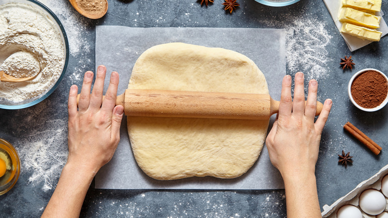 Hands using rolling pin to roll out pastry dough next to baking ingredients like flour and spices