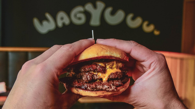 two hands hold a juicy cheeseburger with a back ground that says "waygyuuuu."