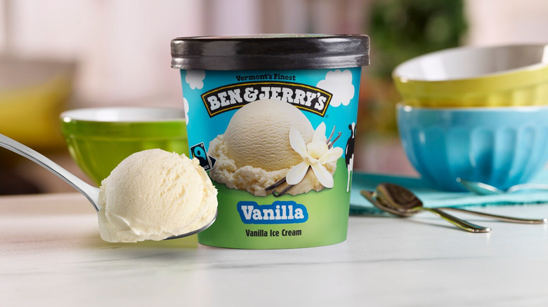 A pint of Benn & Jerry's ice cream next to a spoon filled with the product. In the background there are green, yellow and blue bowls as well as more spoons.
