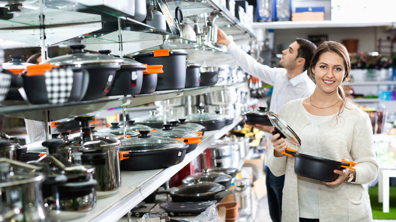 A woman standing next to a shelf filled with different pots and pans, holding a pan and lid and looking at the camera.