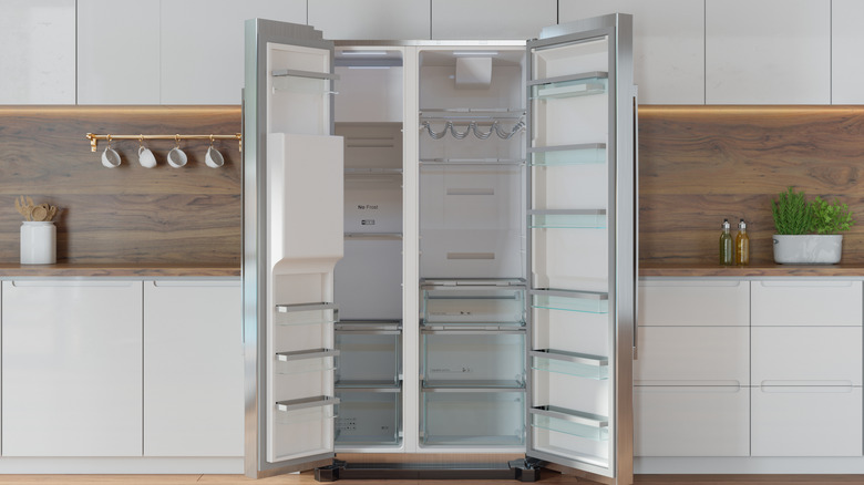An open, empty fridge, standing in the center of a modern kitchen.