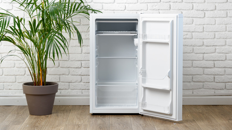 Miniature white standalone freezer with door open next to potted plant against white brick wall and wooden floor