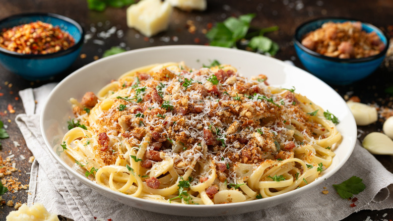 Toasted breadcrumbs on spaghetti in a white bowl on a linen towel and garnishes behind.