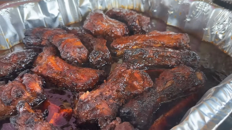 An aluminum tray of cooked party ribs sitting in a pool of dark BBQ sauce