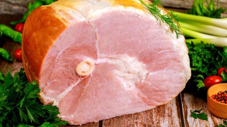 Half bone-in ham on wooden surface next to vegetables like leeks, tomatoes, and herbs