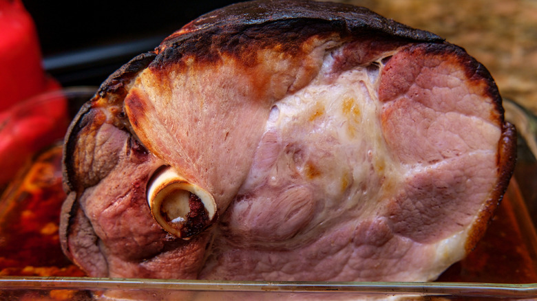 Cooked half bone-in ham with dark smoked exterior in glass pan