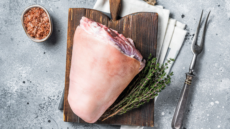 Whole raw ham shank on dark wooden board next to thyme, carving fork, and dish of pink salt