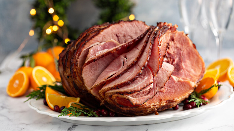 Roasted spiral cut ham on white platter next to orange slices and herbs