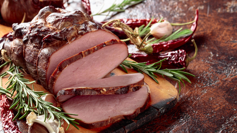 Roasted country ham partially sliced on wooden board next to rosemary and chili peppers